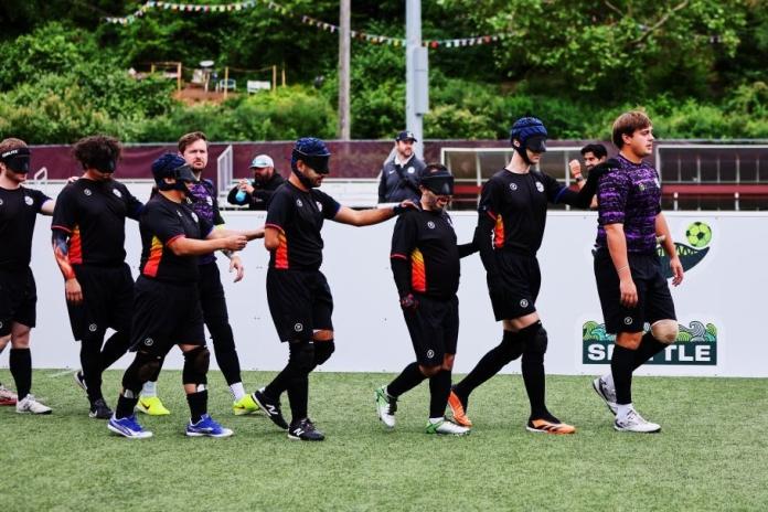 A group of blind football players wearing black uniforms and eye shades walk in a line across an artificial-turf soccer field, with each player resting a hand on the shoulder of the person in front. A player in a purple jersey leads the group.